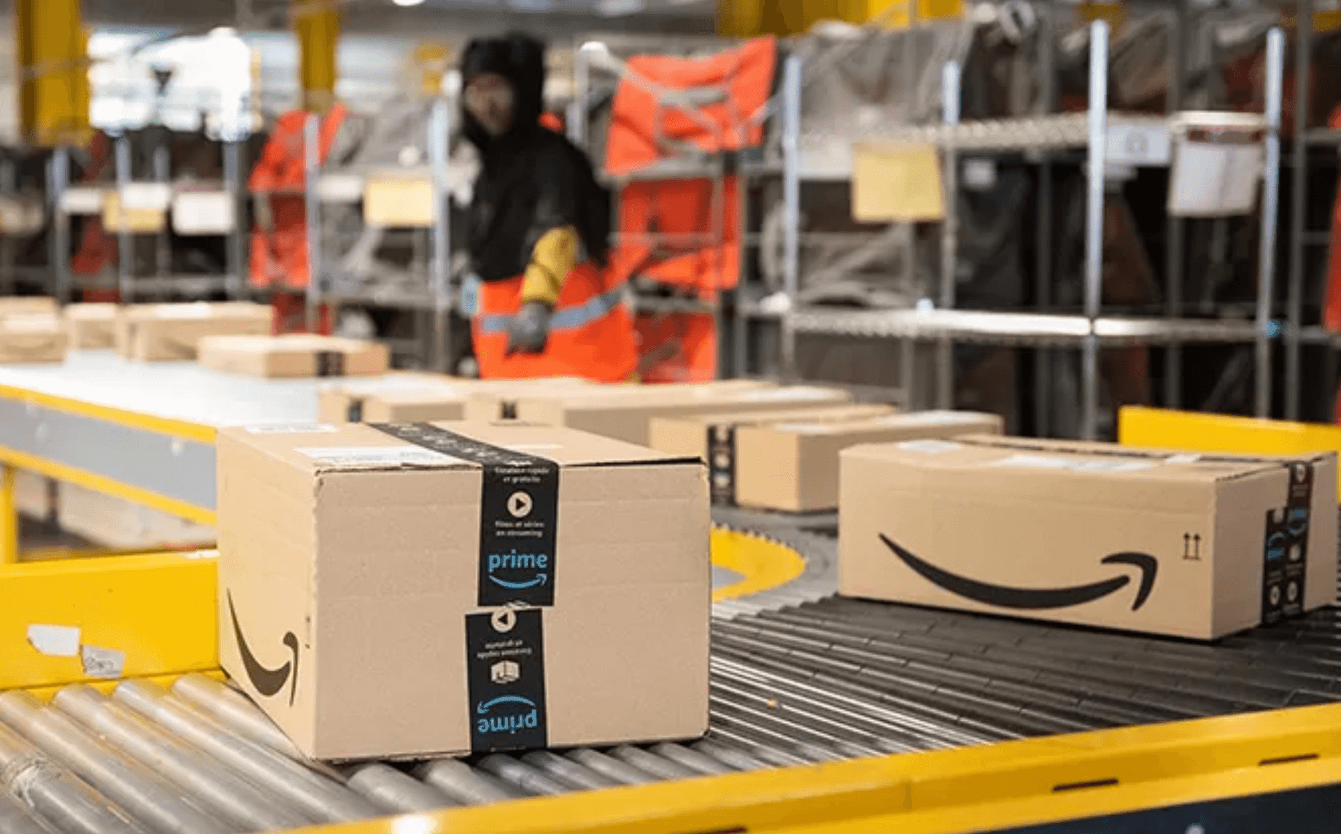 Amazon packages on a conveyor belt inside a warehouse with a worker in safety gear in the background.